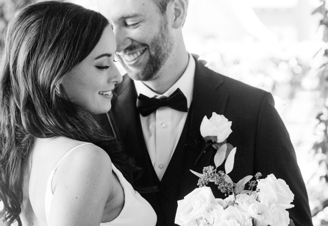 A groom smiles at his bride at an upscale wedding as the bride holds a bouquet of flowers.