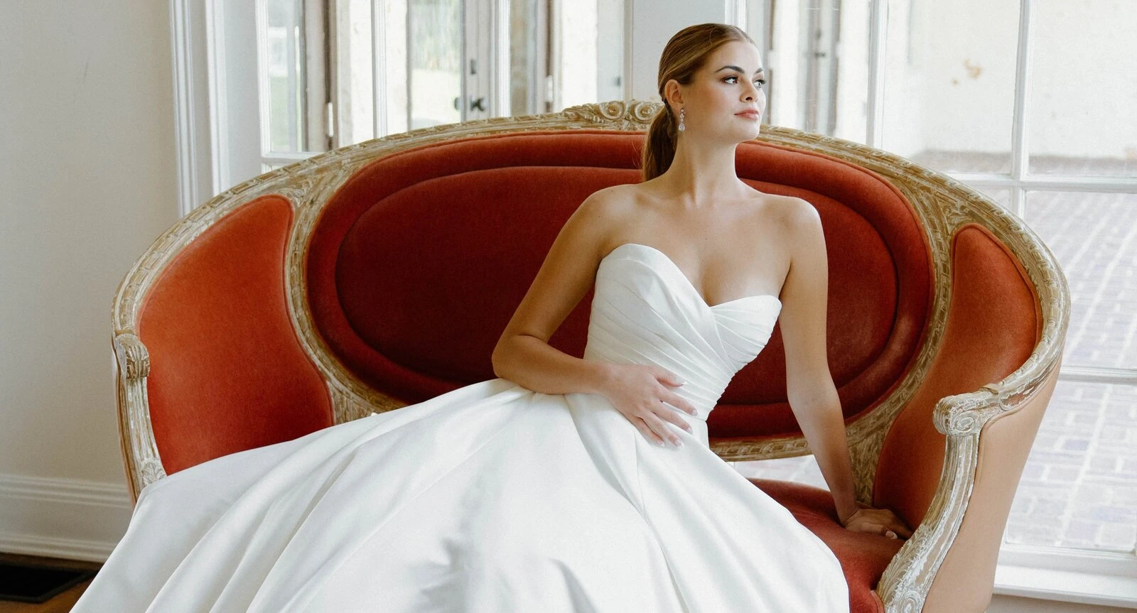 A model wears an Angel Rivera dress while sitting on a red couch