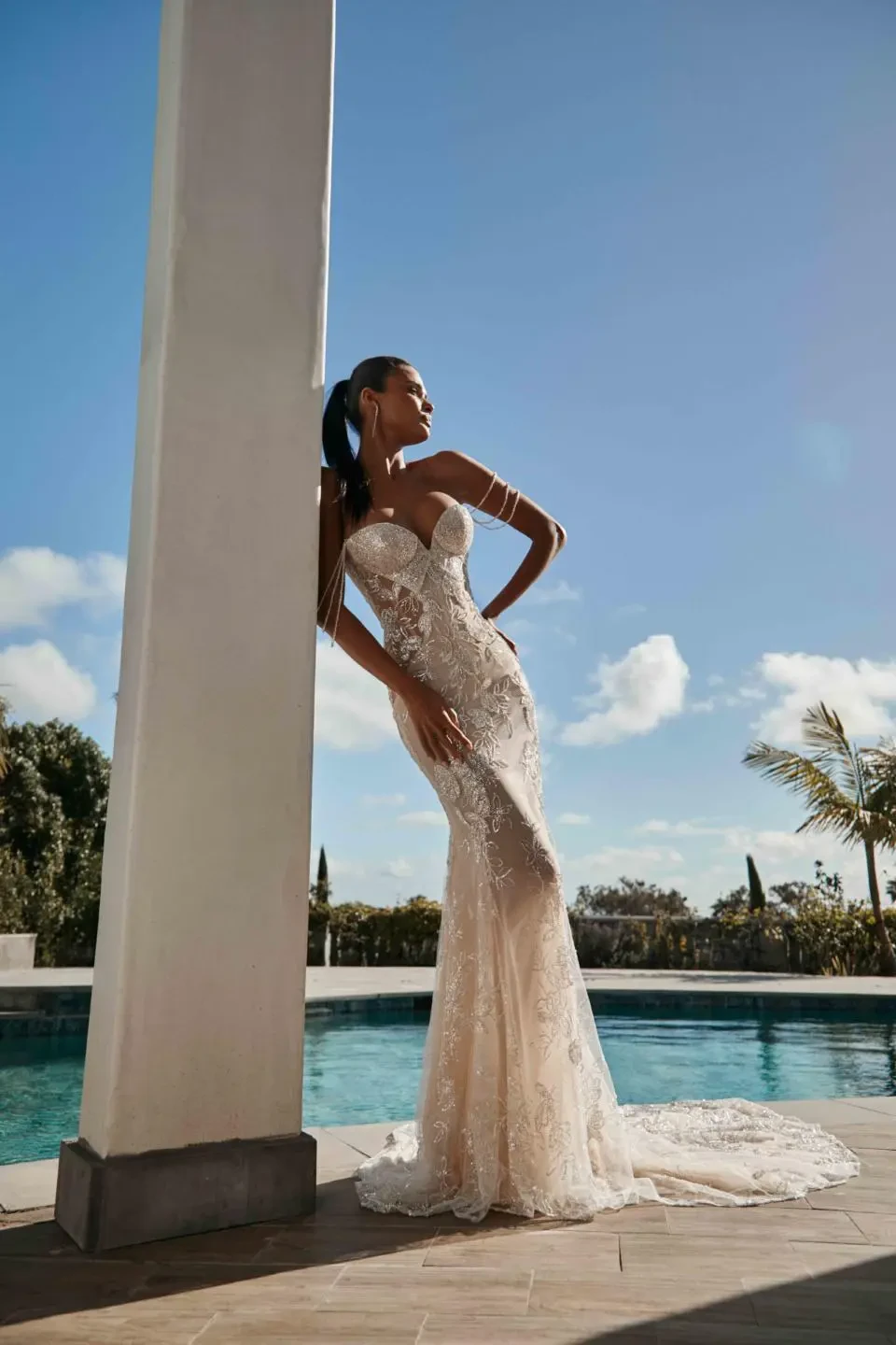 A model wears the Galia Lahav Nala dress outside while leaning against a pillar near a pool