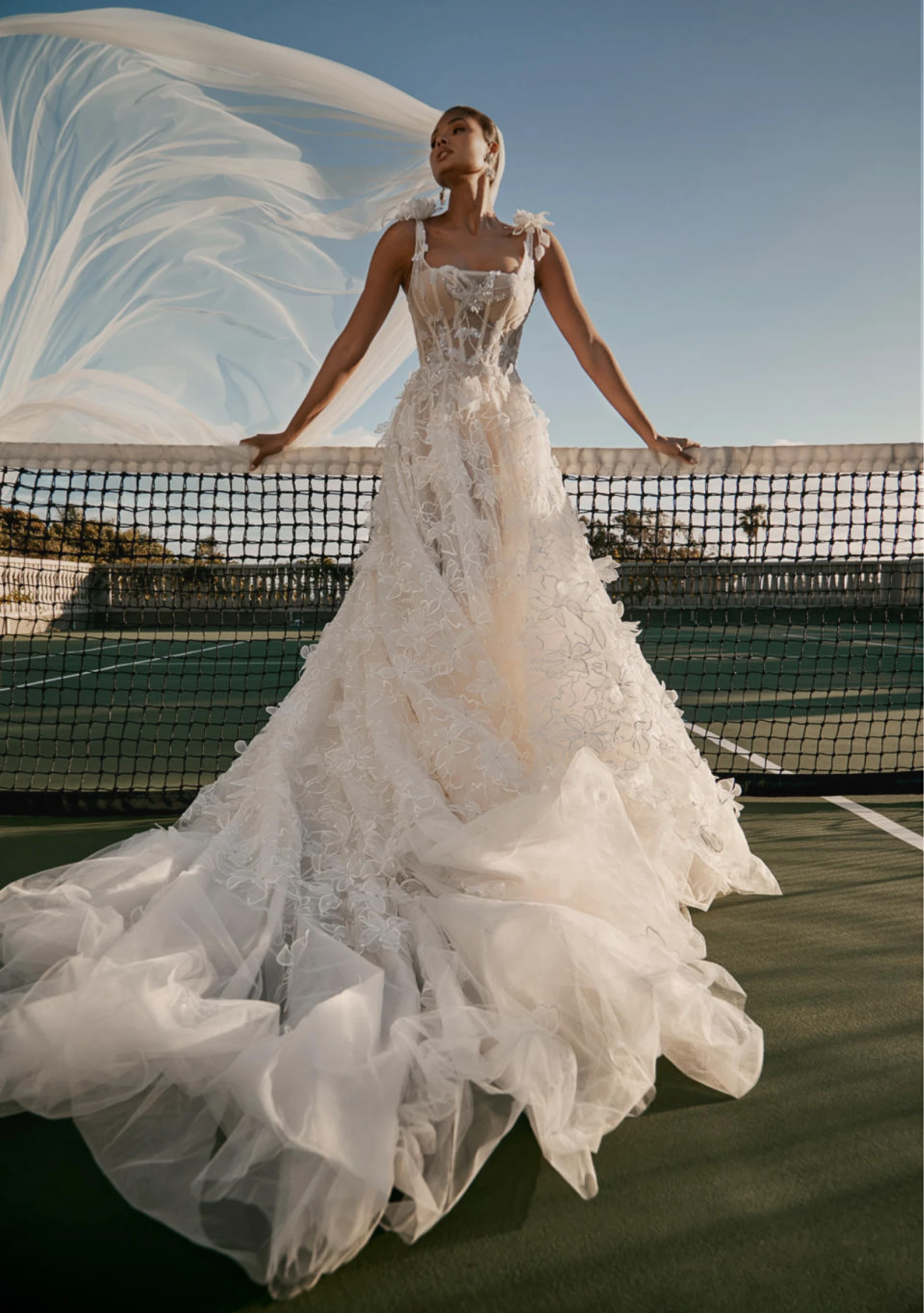 A model wears the Galia Lahav Nirvana dress while leaning against the net of a tennis court